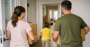 Eine glückliche junge Familie trägt volle Umzugskartons in die neue Wohnung. Sie gehen durch die Haustür ins Haus. Die Familie ist nur von hinten zu sehen. Das Kind rennt spielerisch vor den Eltern ins Haus. Förderung Familien